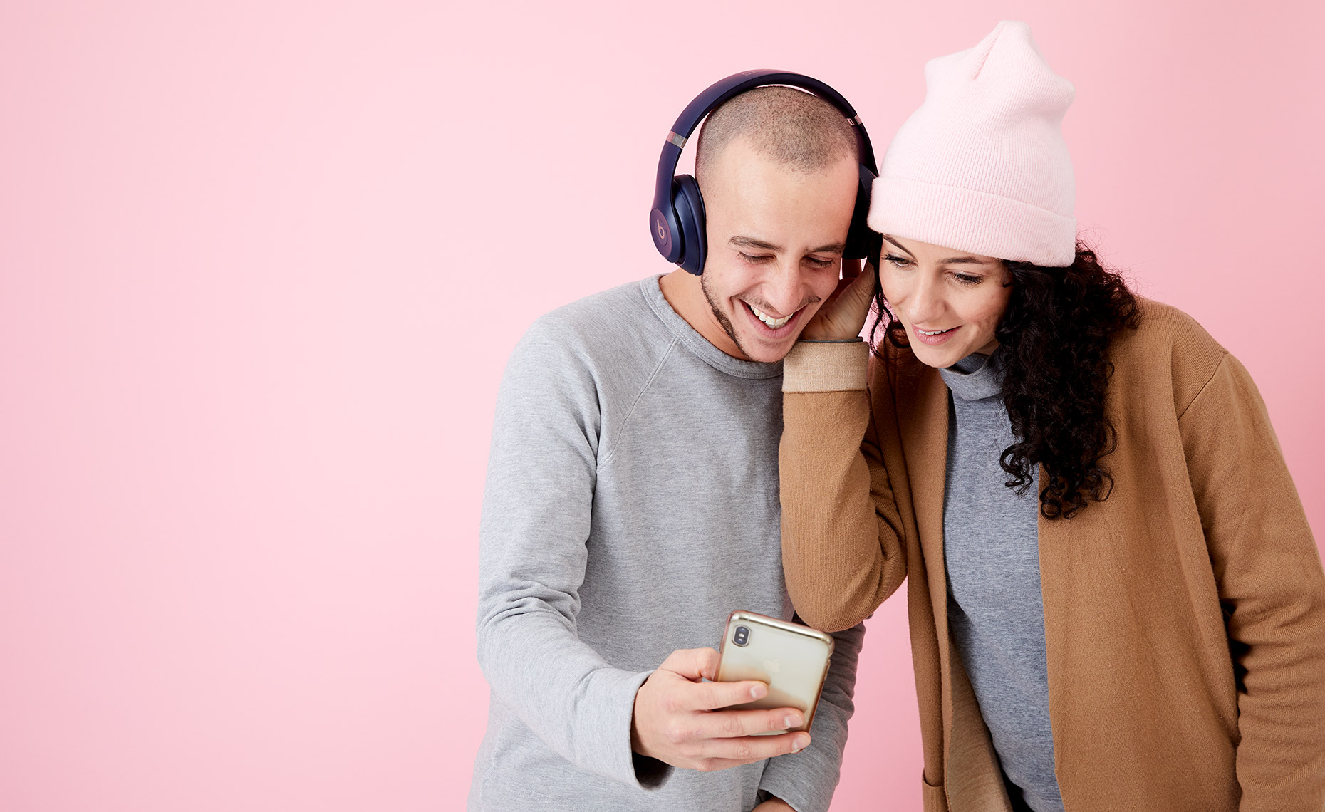 Two young attractive friends leaning in to look at a smartphone screen together, sharing headphones. Photographed against a bright pink background in a studio in New York.