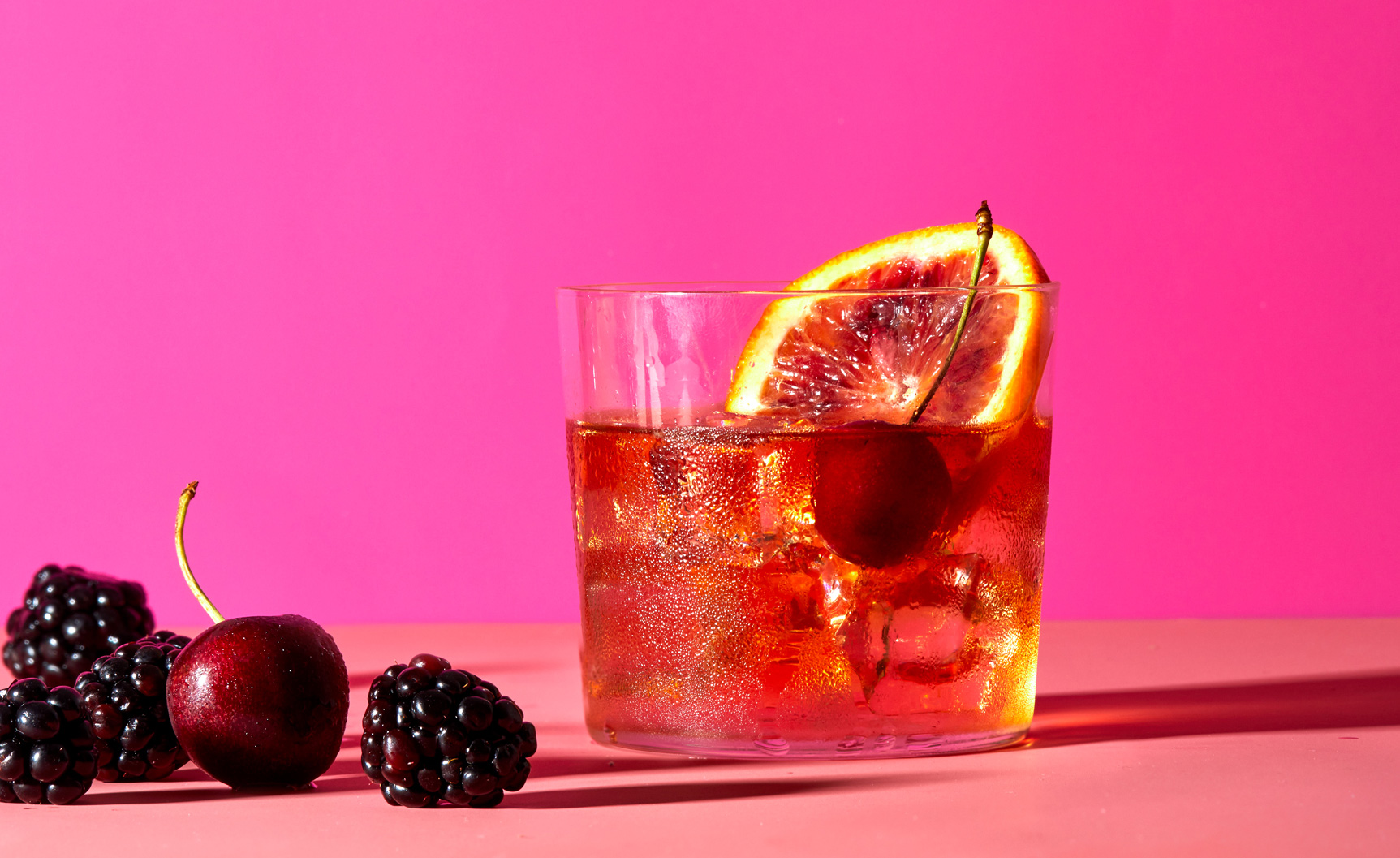 Whiskey cocktail in front of bright colorful pink background with fresh berries garnish.